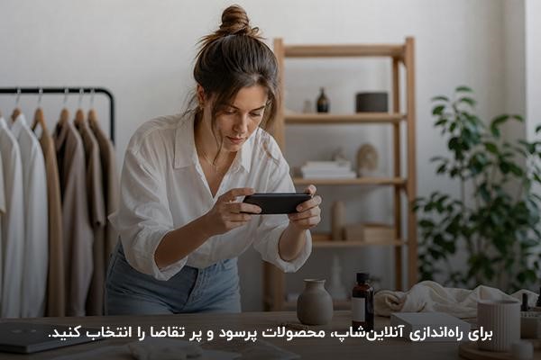 زنی جوان در حال عکس گرفتن از محصول روی میز چوبی با پسزمینه لباسهای آویزان و میز چوبی