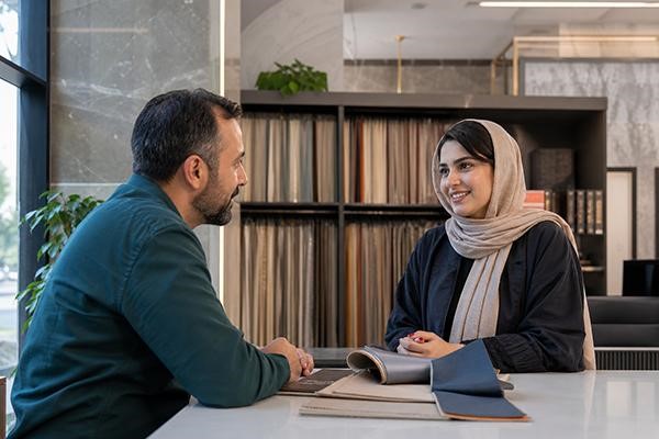 زنی با لباس مشکی و شال کرم در حال صحبت با فروشنده مرد با لباس آبی در فضای داخلی مغازه و انتخاب پارچه